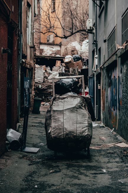 A person wearing a yellow and grey checkered shirt, grey trousers, and bright green gloves is holding open a large black rubbish bag, ready for waste disposal. The bag is made of shiny plastic materia