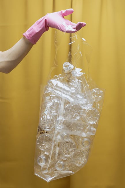 A person's arm wearing a pink rubber glove holds a clear plastic bag filled with various crumpled and flattened transparent plastic bottles and containers. The bag appears to be ready for disposal or 